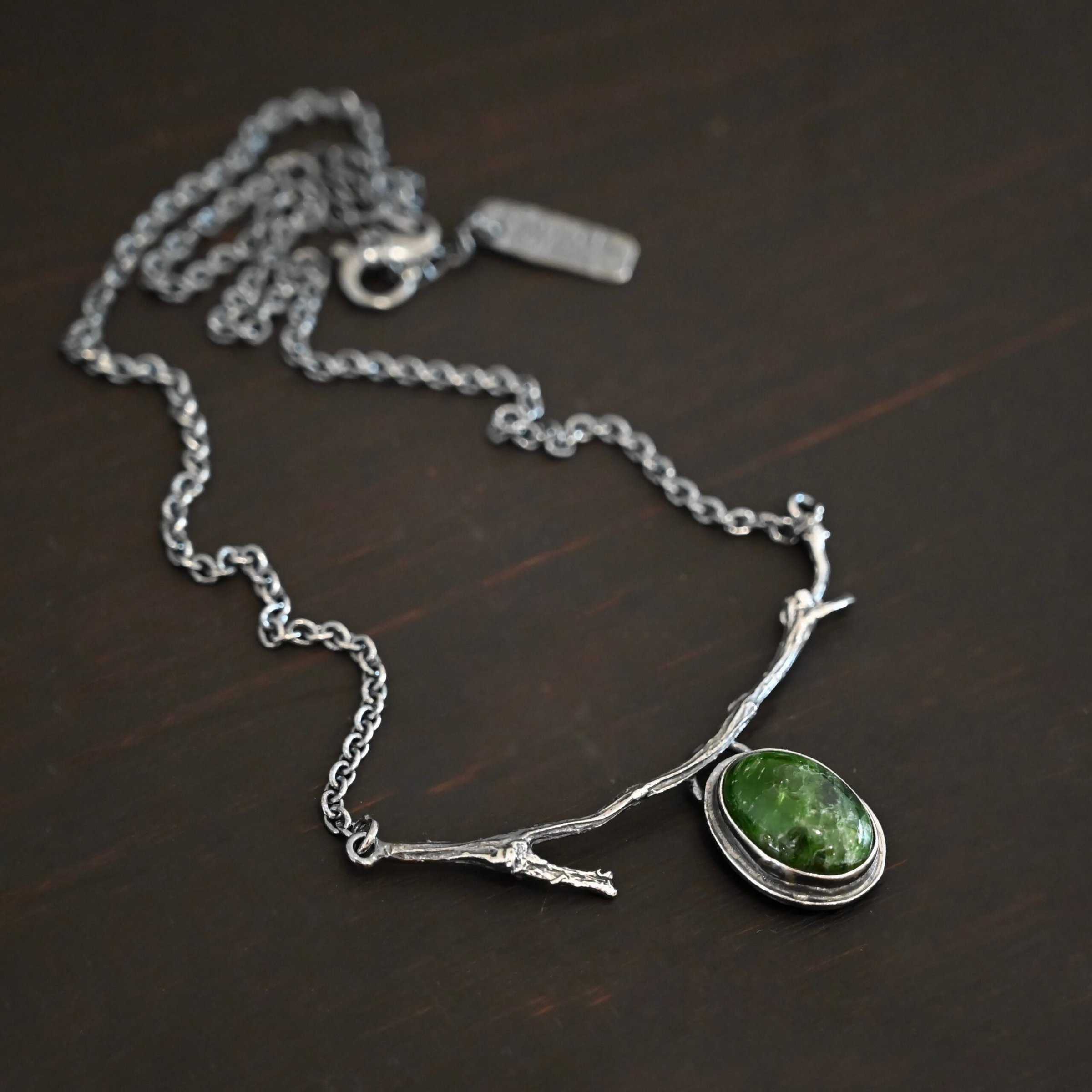 Endless Forest Necklace Close up of the "Endless Forest" Necklace. Made out of sterling silver and chrome diopside. The chrome diopside is an oval shape. It is a dark green color. There is a loop on the back of the pendant that is soldered to a casted "twig" that is curved. Each end of the twig has a loop that a chain is attached to. Each chain leads back to a clasp to connect the necklace. Necklace is shown against a dark background.