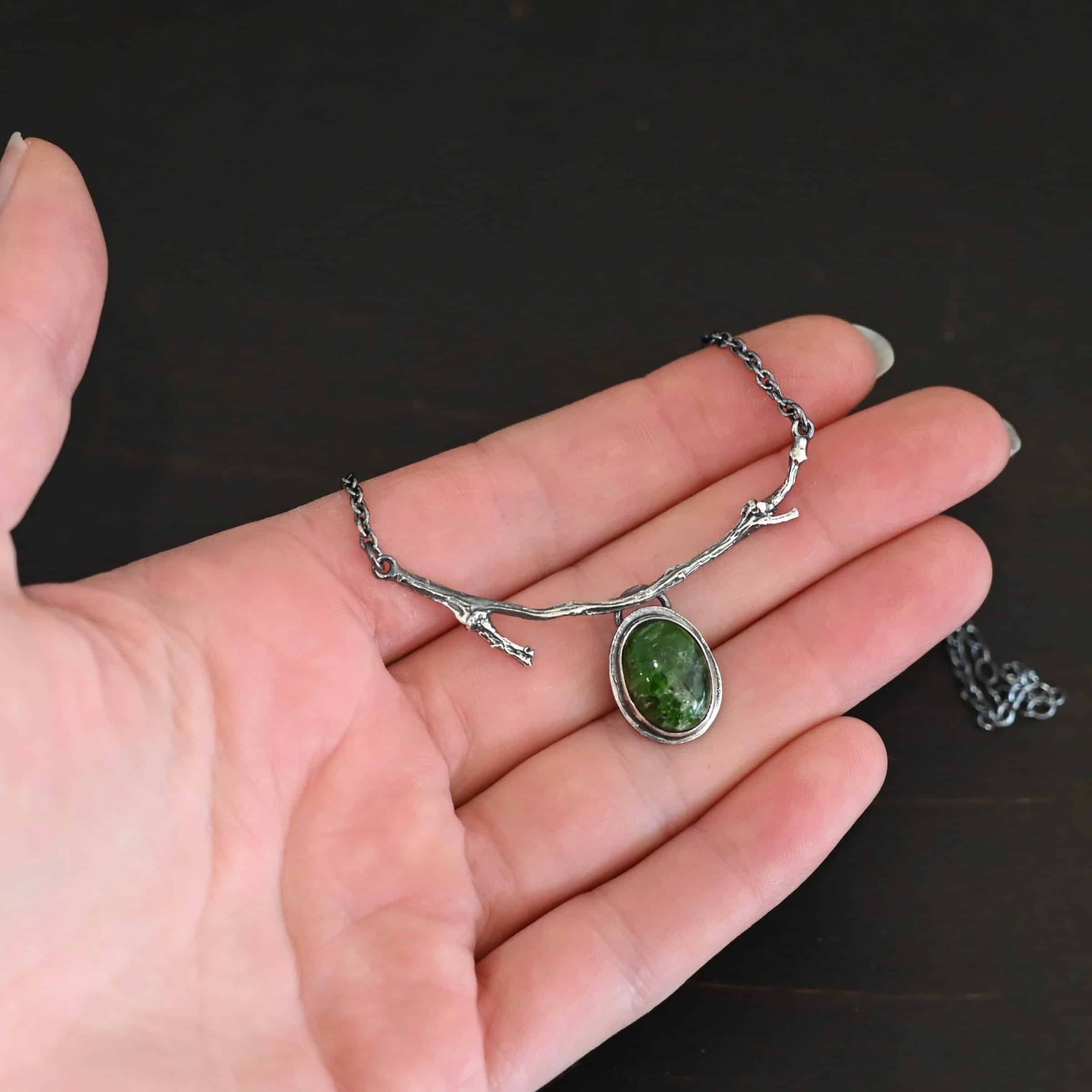Endless Forest Necklace Close up of the "Endless Forest" Necklace. Made out of sterling silver and chrome diopside. The chrome diopside is an oval shape. It is a dark green color. There is a loop on the back of the pendant that is soldered to a casted "twig" that is curved. Each end of the twig has a loop that a chain is attached to. Each chain leads back to a clasp to connect the necklace. Necklace is shown on the fingertips of a hand to demonstrate size.