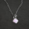 Close up of the "Betty" Necklace. Made out of sterling silver and kunzite. There is a diamond shaped, light pink kunzite in the center. The rest of the setting around the kunzite is hammered sterling silver. There is a loop on the back of the pendant and there are jump rings hanging from the hoop to attach it to the chain for the necklace. Necklace is laying against a dark background.