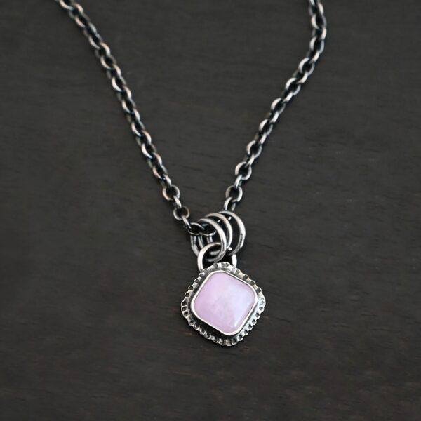 Close up of the "Betty" Necklace. Made out of sterling silver and kunzite. There is a diamond shaped, light pink kunzite in the center. The rest of the setting around the kunzite is hammered sterling silver. There is a loop on the back of the pendant and there are jump rings hanging from the hoop to attach it to the chain for the necklace. Necklace is laying against a dark background.