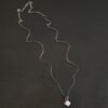 Close up of the "Betty" Necklace. Made out of sterling silver and kunzite. There is a diamond shaped, light pink kunzite in the center. The rest of the setting around the kunzite is hammered sterling silver. There is a loop on the back of the pendant and there are jump rings hanging from the hoop to attach it to the chain for the necklace. Necklace is laying against a dark background.