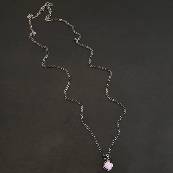 Close up of the "Betty" Necklace. Made out of sterling silver and kunzite. There is a diamond shaped, light pink kunzite in the center. The rest of the setting around the kunzite is hammered sterling silver. There is a loop on the back of the pendant and there are jump rings hanging from the hoop to attach it to the chain for the necklace. Necklace is laying against a dark background.