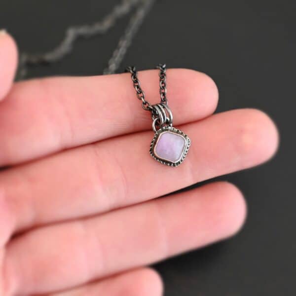 Close up of the "Betty" Necklace. Made out of sterling silver and kunzite. There is a diamond shaped, light pink kunzite in the center. The rest of the setting around the kunzite is hammered sterling silver. There is a loop on the back of the pendant and there are jump rings hanging from the hoop to attach it to the chain for the necklace. Necklace is laying against the fingertips of a hand to demonstrate size of the pendant.