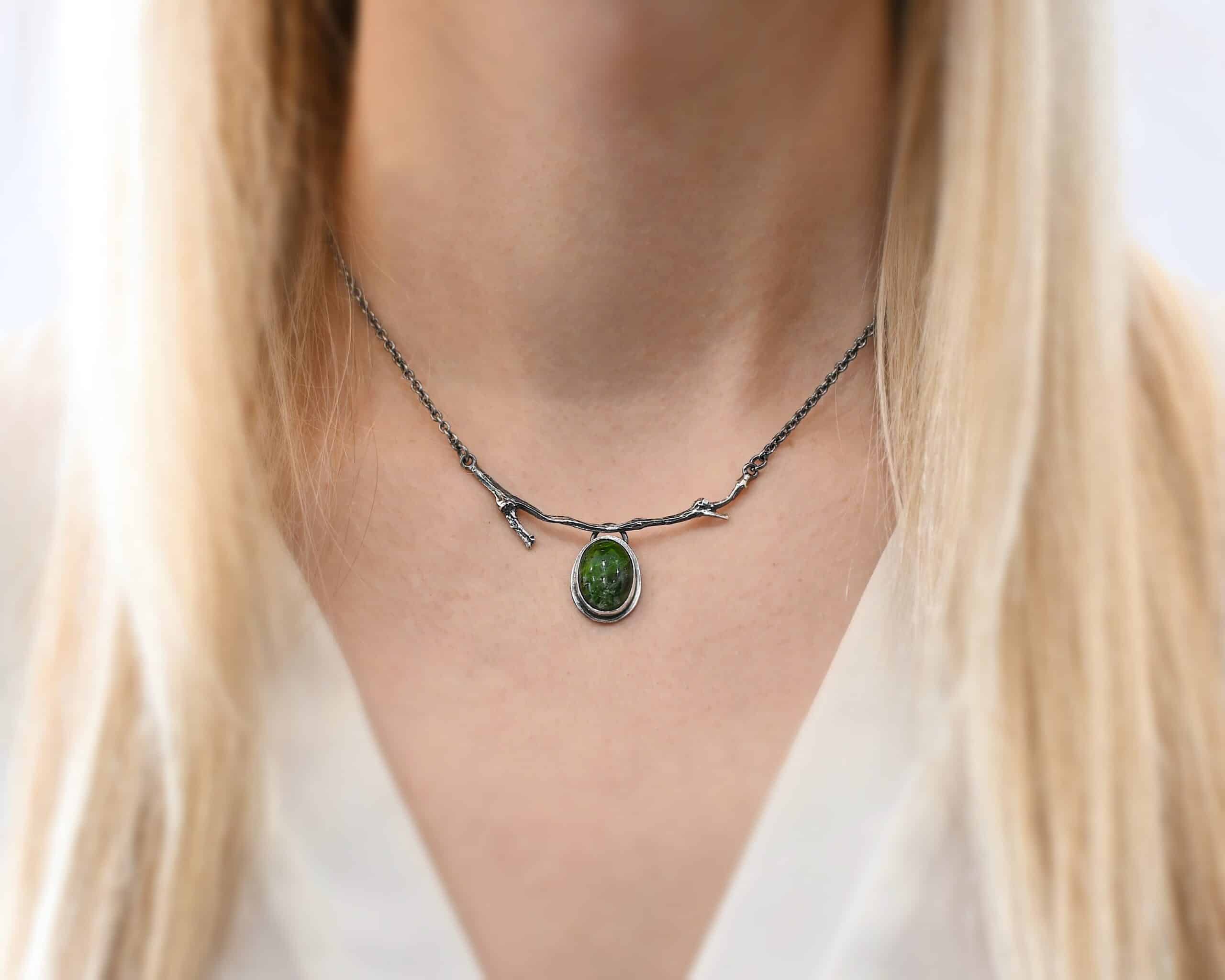 Endless Forest Necklace Close up of the "Endless Forest" Necklace. Made out of sterling silver and chrome diopside. The chrome diopside is an oval shape. It is a dark green color. There is a loop on the back of the pendant that is soldered to a casted "twig" that is curved. Each end of the twig has a loop that a chain is attached to. Each chain leads back to a clasp to connect the necklace. Necklace is worn at 16" long and model is wearing a white v-neck shirt.