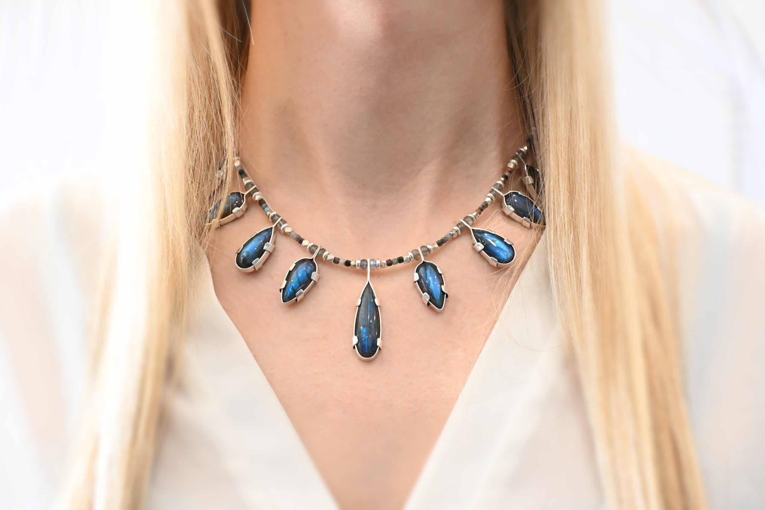 Rain Dance Necklace Close up of the "Rain Dance" Necklace. Made out of sterling silver and labradorite, with labradorite beads. There are eight teardrop shaped labradorites, and each stone alternates from point facing upwards to point facing downwards. Each one is connected to a chain that is made out of labradorite and chrysocolla beads. Each labradorite has four tabs, two on each side of the stone, that are clamped down on the edges of it to keep it held in. Necklace is worn at 16" long and model is wearing a white v-neck shirt.
