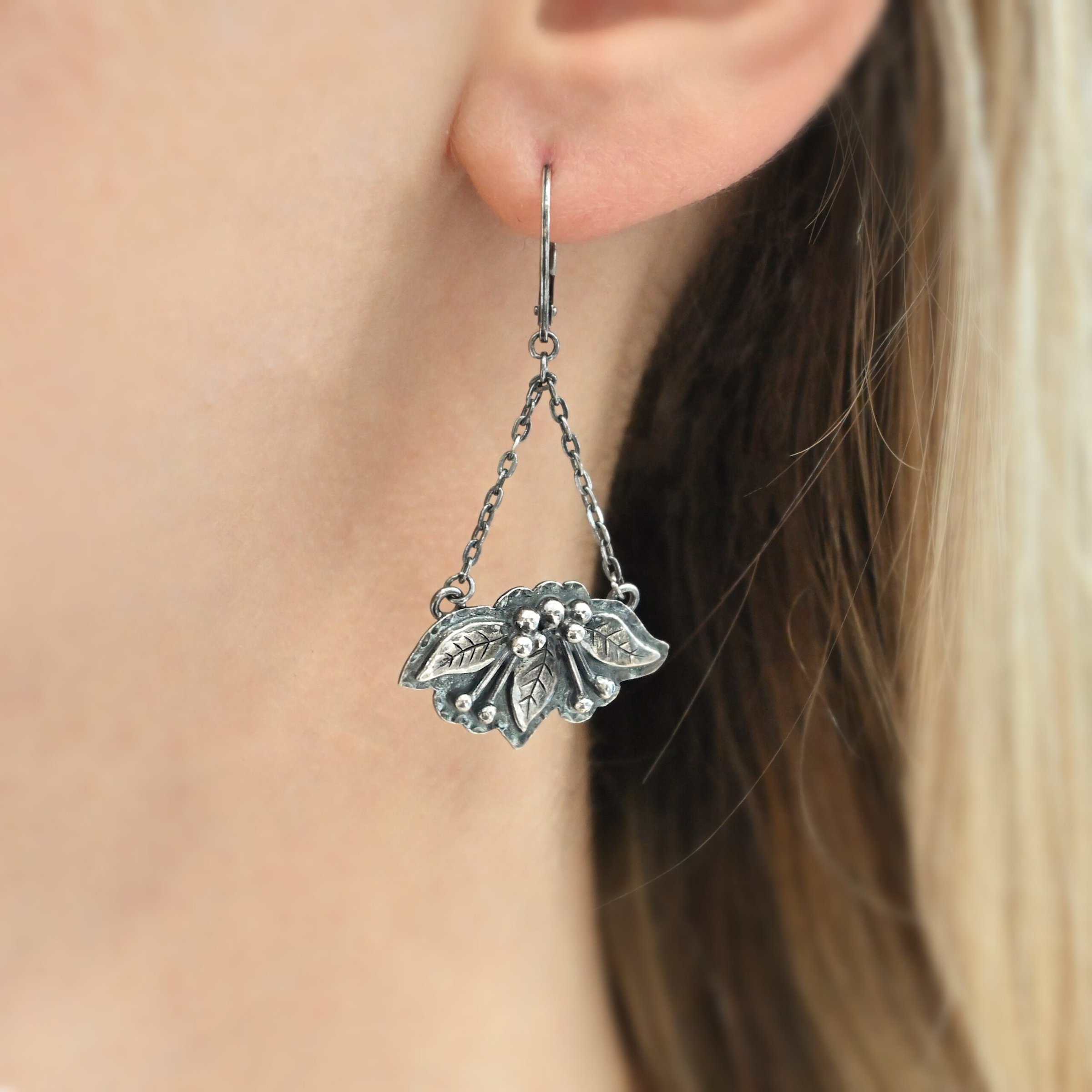 "Paisley" Earring shown on a model. There is a piece of silver with three silver leaves on it, along with silver ball detail and small sticks of silver. It looks like a flower. There are two small strands of silver on the top that create a triangle with a hollow center. There is an ear wire at the top that connects the two strands of silver to create the triangle shape.