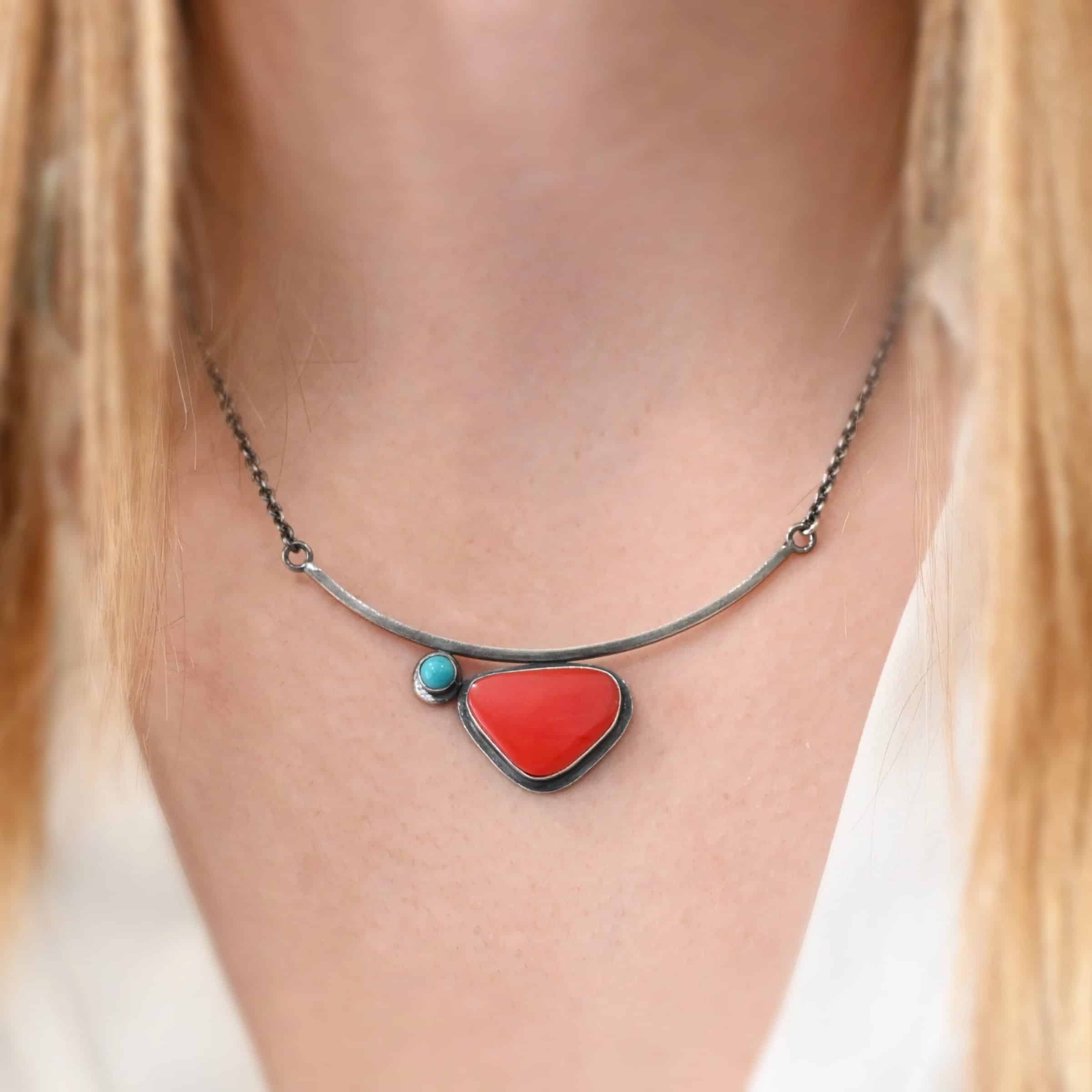 Sienna Necklace New Close up of the "Sienna" Necklace. Made out of sterling silver, rosarita, and turquoise. The rosarita is a triangle shape and a bright red color. There is a small turquoise dot on the top left corner of the necklace. Both of these are attached to a bar pendant that is curved. On both ends of the bar pendant are loops from which the chain attaches to. Model is wearing the necklace at 18" long and model is wearing a white v-neck shirt.
