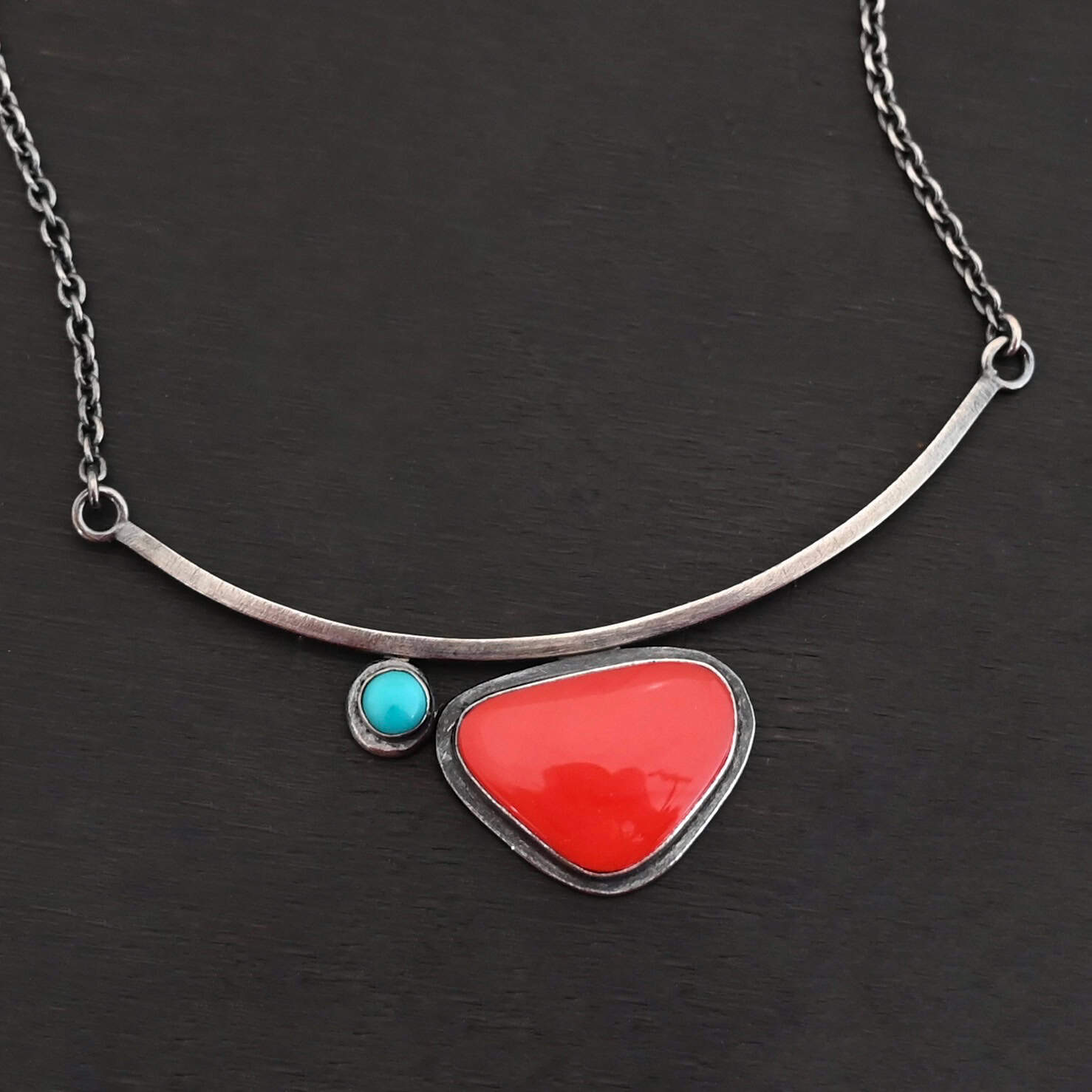 Sienna Necklace New Close up of the "Sienna" Necklace. Made out of sterling silver, rosarita, and turquoise. The rosarita is a triangle shape and a bright red color. There is a small turquoise dot on the top left corner of the necklace. Both of these are attached to a bar pendant that is curved. On both ends of the bar pendant are loops from which the chain attaches to. Necklace is laying against a dark background.