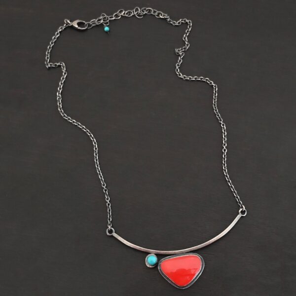 Close up of the "Sienna" Necklace. Made out of sterling silver, rosarita, and turquoise. The rosarita is a triangle shape and a bright red color. There is a small turquoise dot on the top left corner of the necklace. Both of these are attached to a bar pendant that is curved. On both ends of the bar pendant are loops from which the chain attaches to. Necklace is laying against a dark background.