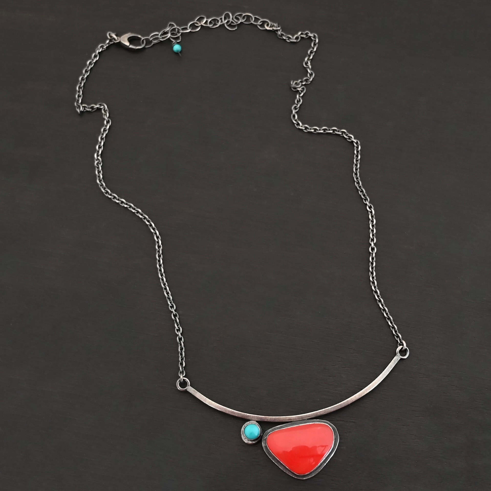 Sienna Necklace New Close up of the "Sienna" Necklace. Made out of sterling silver, rosarita, and turquoise. The rosarita is a triangle shape and a bright red color. There is a small turquoise dot on the top left corner of the necklace. Both of these are attached to a bar pendant that is curved. On both ends of the bar pendant are loops from which the chain attaches to. Necklace is laying against a dark background.