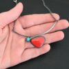 Close up of the "Sienna" Necklace. Made out of sterling silver, rosarita, and turquoise. The rosarita is a triangle shape and a bright red color. There is a small turquoise dot on the top left corner of the necklace. Both of these are attached to a bar pendant that is curved. On both ends of the bar pendant are loops from which the chain attaches to. Necklace is laying against the palm of a hand to demonstrate size.