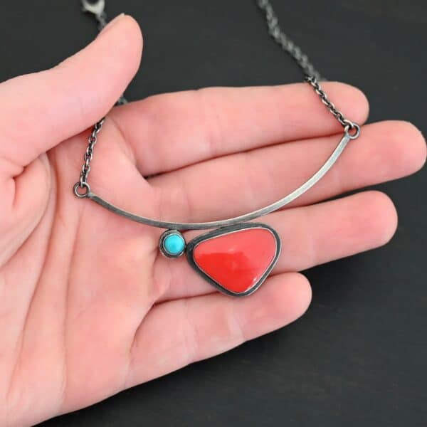 Close up of the "Sienna" Necklace. Made out of sterling silver, rosarita, and turquoise. The rosarita is a triangle shape and a bright red color. There is a small turquoise dot on the top left corner of the necklace. Both of these are attached to a bar pendant that is curved. On both ends of the bar pendant are loops from which the chain attaches to. Necklace is laying against the palm of a hand to demonstrate size.