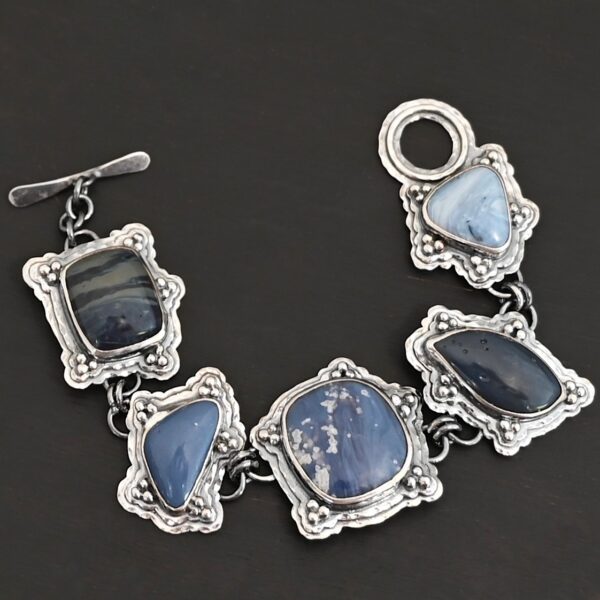 Close up of the "Frozen Lake" Bracelet. Made out of sterling silver & leland blue. There are five leland blue stones, each a different shape and color of blue. There is a toggle clasp on the back of the bracelet to connect each end. Each stone has fifteen sterling silver balls that are in groups of three circling around the stone. Bracelet is laying against a dark background.