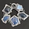 Close up of the "Frozen Lake" Bracelet. Made out of sterling silver & leland blue. There are five leland blue stones, each a different shape and color of blue. There is a toggle clasp on the back of the bracelet to connect each end. Each stone has fifteen sterling silver balls that are in groups of three circling around the stone. Bracelet is laying against a dark background.