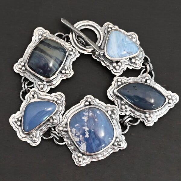 Close up of the "Frozen Lake" Bracelet. Made out of sterling silver & leland blue. There are five leland blue stones, each a different shape and color of blue. There is a toggle clasp on the back of the bracelet to connect each end. Each stone has fifteen sterling silver balls that are in groups of three circling around the stone. Bracelet is laying against a dark background.