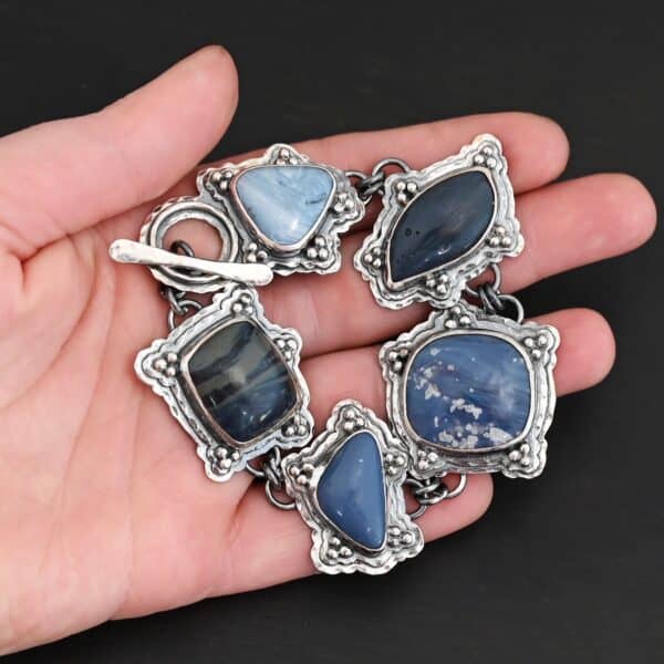 Close up of the "Frozen Lake" Bracelet. Made out of sterling silver & leland blue. There are five leland blue stones, each a different shape and color of blue. There is a toggle clasp on the back of the bracelet to connect each end. Each stone has fifteen sterling silver balls that are in groups of three circling around the stone. Bracelet is lying against the fingertips of a hand.