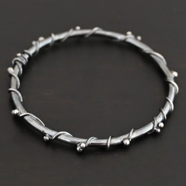 Close up of the "Ice Berry" Bangle. Made out of sterling silver and oxidized. Bangle has sterling silver "vines" wrapped all the way around it with small silver balls that are scattered around the bangle and vines. Bangle is laying against a dark background.