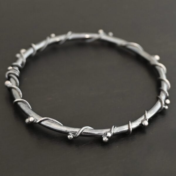 Close up of the "Ice Berry" Bangle. Made out of sterling silver and oxidized. Bangle has sterling silver "vines" wrapped all the way around it with small silver balls that are scattered around the bangle and vines. Bangle is laying against a dark background.