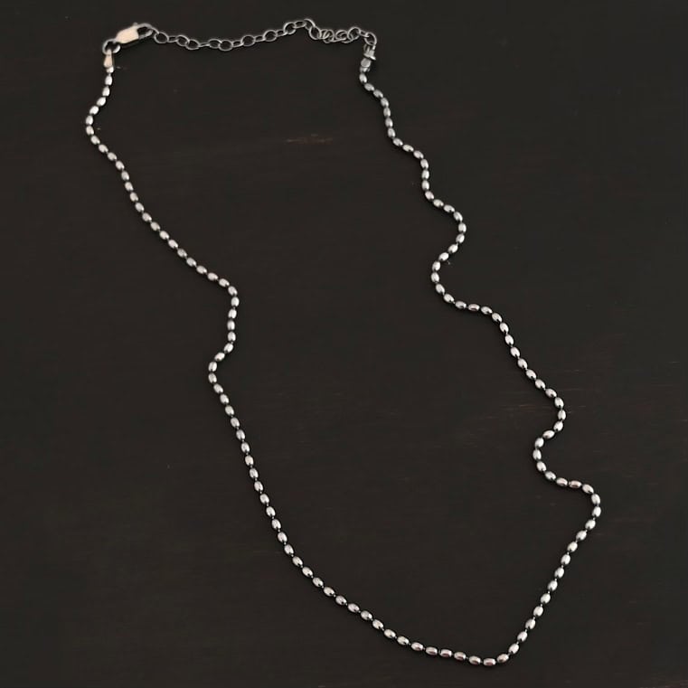 "Bryn Beaded" Chain as shown up close. Chain is shown as oxidized with 1.5mm oval beads that have very little space between them. Chain is shown against a dark background. Chain has a lobster claw clasp and a three inch extension made out of sterling silver.