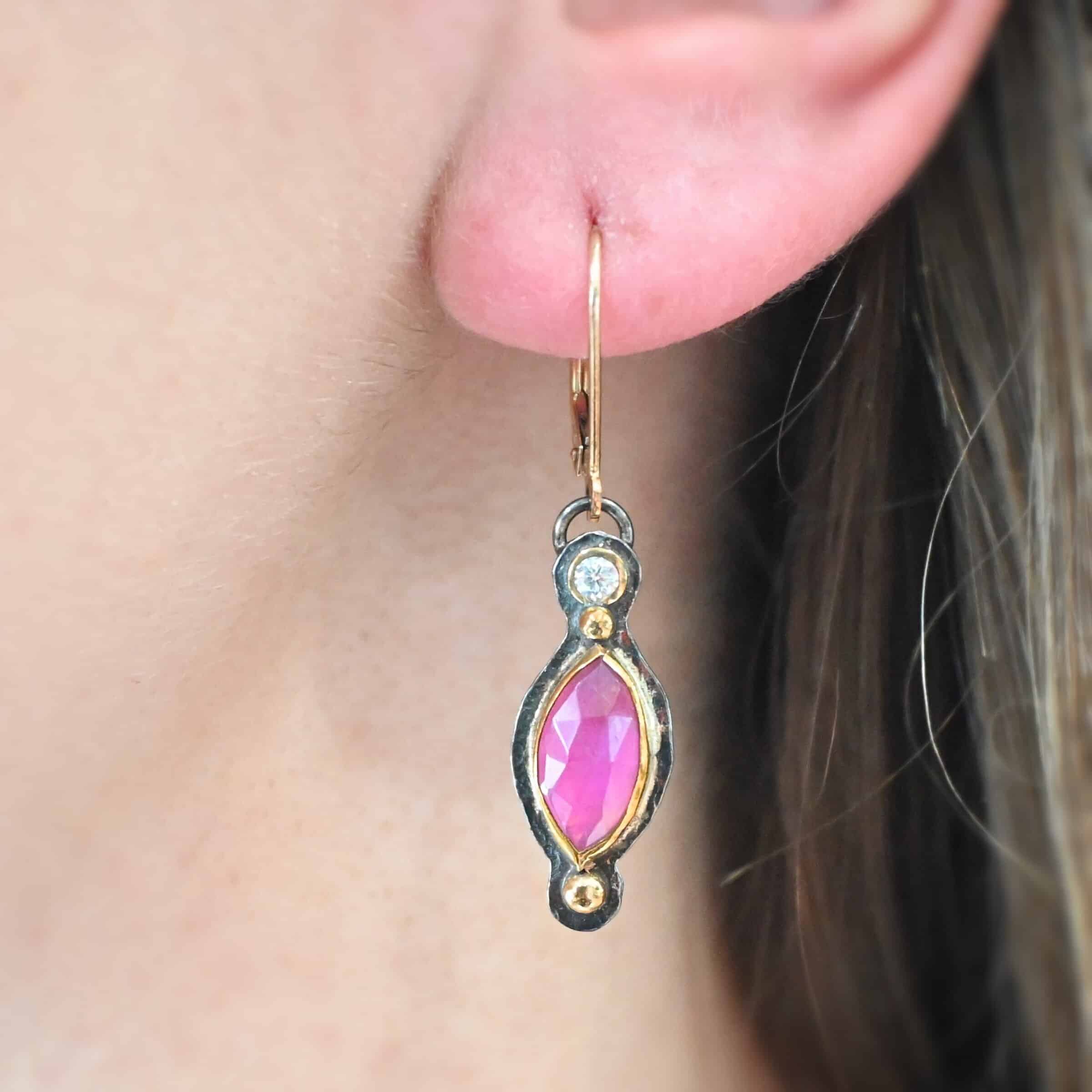 Close up of our "Pink Skies" Earrings. Made out of sterling silver and 18k gold, rubies and moissanites. There are two ruby stones and two moissanites. Both ruby stones are oval shaped with points on each end. Both rubies are set in gold. The left earring has a moissanite set in gold on the bottom of it. The right earring has the moissanite on the top of the earring. Both earrings have two small gold dots on them, one on the top of the earring and one on the bottom. Earrings are on gold leverback ear wires. Earrings are worn by a model up close.