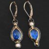 Close up of our "Beyond The Ocean" Earrings. Made out of sterling silver and 18k gold, kyanite and moissanites. There are two kyanite stones and two moissanites. Both kyanite stones are teardrops. One is facing with the point upwards. The other has the point facing downwards. Both kyanites are set in gold. The earring with the point facing upwards has a moissanite on the bottom of it. The right earring with the point facing downwards has the moissanite on the top of the earring. Both earrings has two small gold dots on them, one on the top of the earring and one on the bottom. Earrings are on gold leverback ear wires.