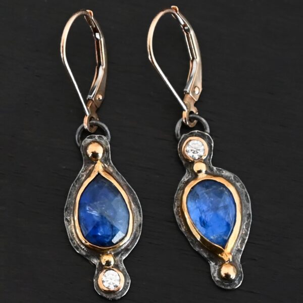 Close up of our "Beyond The Ocean" Earrings. Made out of sterling silver and 18k gold, kyanite and moissanites. There are two kyanite stones and two moissanites. Both kyanite stones are teardrops. One is facing with the point upwards. The other has the point facing downwards. Both kyanites are set in gold. The earring with the point facing upwards has a moissanite on the bottom of it. The right earring with the point facing downwards has the moissanite on the top of the earring. Both earrings has two small gold dots on them, one on the top of the earring and one on the bottom. Earrings are on gold leverback ear wires.
