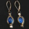 Close up of our "Beyond The Ocean" Earrings. Made out of sterling silver and 18k gold, kyanite and moissanites. There are two kyanite stones and two moissanites. Both kyanite stones are teardrops. One is facing with the point upwards. The other has the point facing downwards. Both kyanites are set in gold. The earring with the point facing upwards has a moissanite on the bottom of it. The right earring with the point facing downwards has the moissanite on the top of the earring. Both earrings has two small gold dots on them, one on the top of the earring and one on the bottom. Earrings are on gold leverback ear wires.