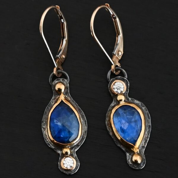 Close up of our "Beyond The Ocean" Earrings. Made out of sterling silver and 18k gold, kyanite and moissanites. There are two kyanite stones and two moissanites. Both kyanite stones are teardrops. One is facing with the point upwards. The other has the point facing downwards. Both kyanites are set in gold. The earring with the point facing upwards has a moissanite on the bottom of it. The right earring with the point facing downwards has the moissanite on the top of the earring. Both earrings has two small gold dots on them, one on the top of the earring and one on the bottom. Earrings are on gold leverback ear wires.