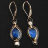 Close up of our "Beyond The Ocean" Earrings. Made out of sterling silver and 18k gold, kyanite and moissanites. There are two kyanite stones and two moissanites. Both kyanite stones are teardrops. One is facing with the point upwards. The other has the point facing downwards. Both kyanites are set in gold. The earring with the point facing upwards has a moissanite on the bottom of it. The right earring with the point facing downwards has the moissanite on the top of the earring. Both earrings has two small gold dots on them, one on the top of the earring and one on the bottom. Earrings are on gold leverback ear wires.