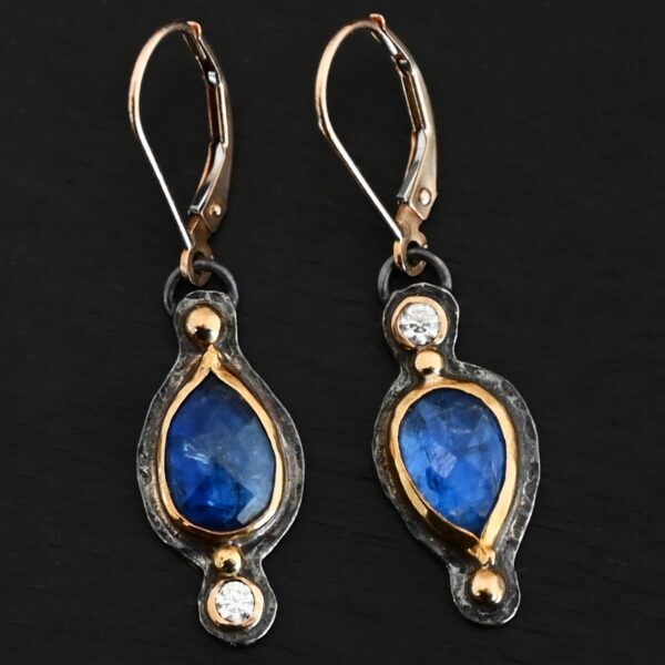 Close up of our "Beyond The Ocean" Earrings. Made out of sterling silver and 18k gold, kyanite and moissanites. There are two kyanite stones and two moissanites. Both kyanite stones are teardrops. One is facing with the point upwards. The other has the point facing downwards. Both kyanites are set in gold. The earring with the point facing upwards has a moissanite on the bottom of it. The right earring with the point facing downwards has the moissanite on the top of the earring. Both earrings has two small gold dots on them, one on the top of the earring and one on the bottom. Earrings are on gold leverback ear wires.