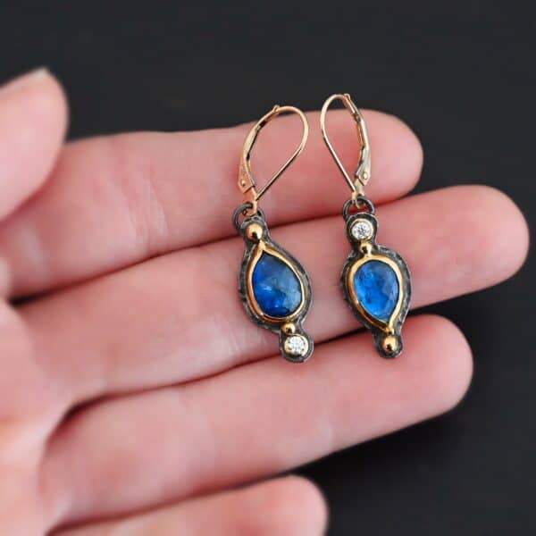 Close up of our "Beyond The Ocean" Earrings. Made out of sterling silver and 18k gold, kyanite and moissanites. There are two kyanite stones and two moissanites. Both kyanite stones are teardrops. One is facing with the point upwards. The other has the point facing downwards. Both kyanites are set in gold. The earring with the point facing upwards has a moissanite on the bottom of it. The right earring with the point facing downwards has the moissanite on the top of the earring. Both earrings has two small gold dots on them, one on the top of the earring and one on the bottom. Earrings are on gold leverback ear wires. Earrings are held by a model up close.