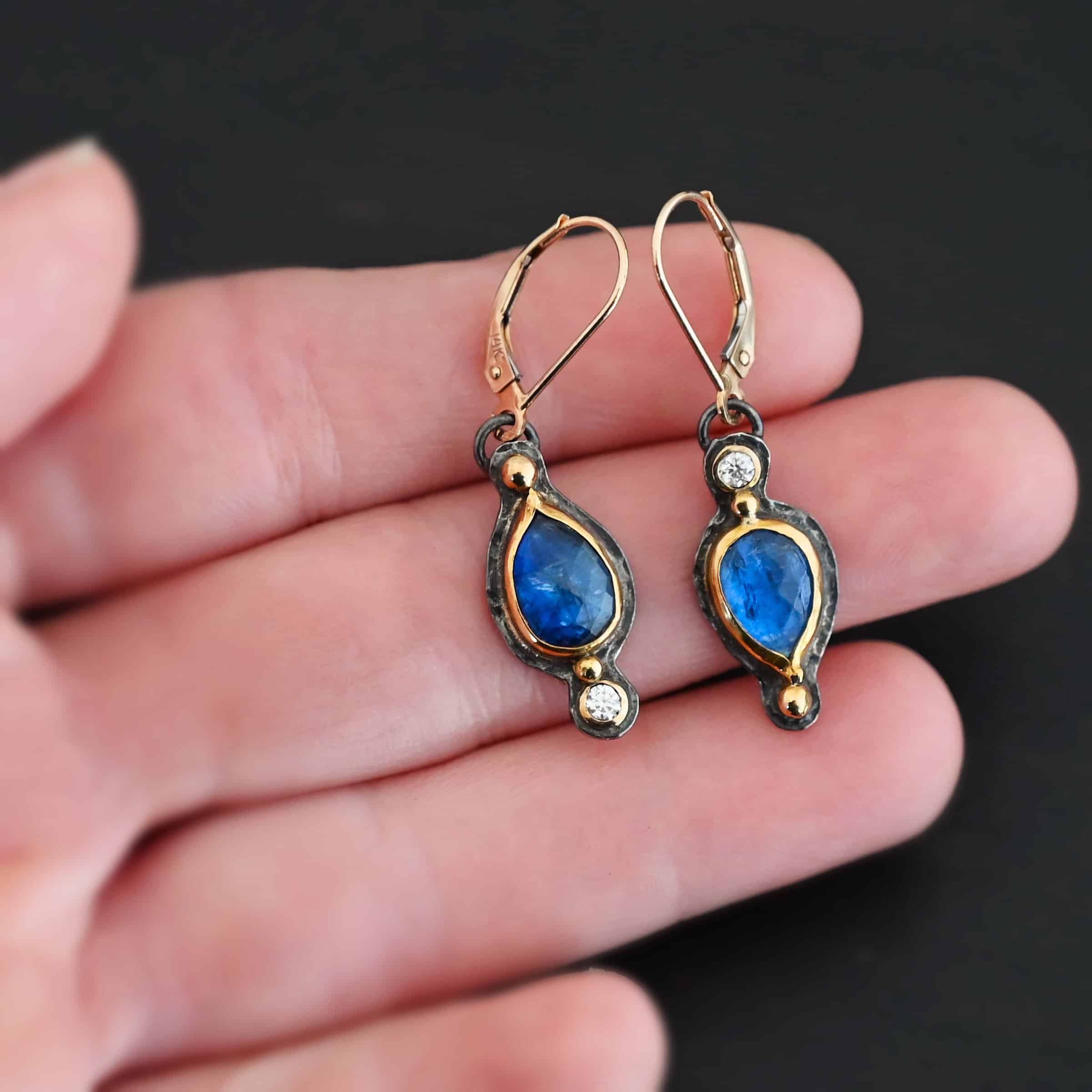 Beyond The Ocean Earrings New Close up of our "Beyond The Ocean" Earrings. Made out of sterling silver and 18k gold, kyanite and moissanites. There are two kyanite stones and two moissanites. Both kyanite stones are teardrops. One is facing with the point upwards. The other has the point facing downwards. Both kyanites are set in gold. The earring with the point facing upwards has a moissanite on the bottom of it. The right earring with the point facing downwards has the moissanite on the top of the earring. Both earrings has two small gold dots on them, one on the top of the earring and one on the bottom. Earrings are on gold leverback ear wires. Earrings are held by a model up close.