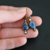 Close up of our "Beyond The Ocean" Earrings. Made out of sterling silver and 18k gold, kyanite and moissanites. There are two kyanite stones and two moissanites. Both kyanite stones are teardrops. One is facing with the point upwards. The other has the point facing downwards. Both kyanites are set in gold. The earring with the point facing upwards has a moissanite on the bottom of it. The right earring with the point facing downwards has the moissanite on the top of the earring. Both earrings has two small gold dots on them, one on the top of the earring and one on the bottom. Earrings are on gold leverback ear wires. Earrings are held by a model up close.