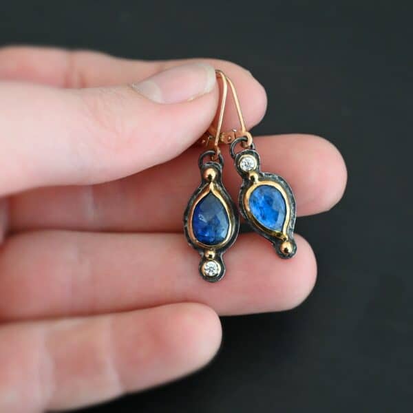 Close up of our "Beyond The Ocean" Earrings. Made out of sterling silver and 18k gold, kyanite and moissanites. There are two kyanite stones and two moissanites. Both kyanite stones are teardrops. One is facing with the point upwards. The other has the point facing downwards. Both kyanites are set in gold. The earring with the point facing upwards has a moissanite on the bottom of it. The right earring with the point facing downwards has the moissanite on the top of the earring. Both earrings has two small gold dots on them, one on the top of the earring and one on the bottom. Earrings are on gold leverback ear wires. Earrings are held by a model up close.