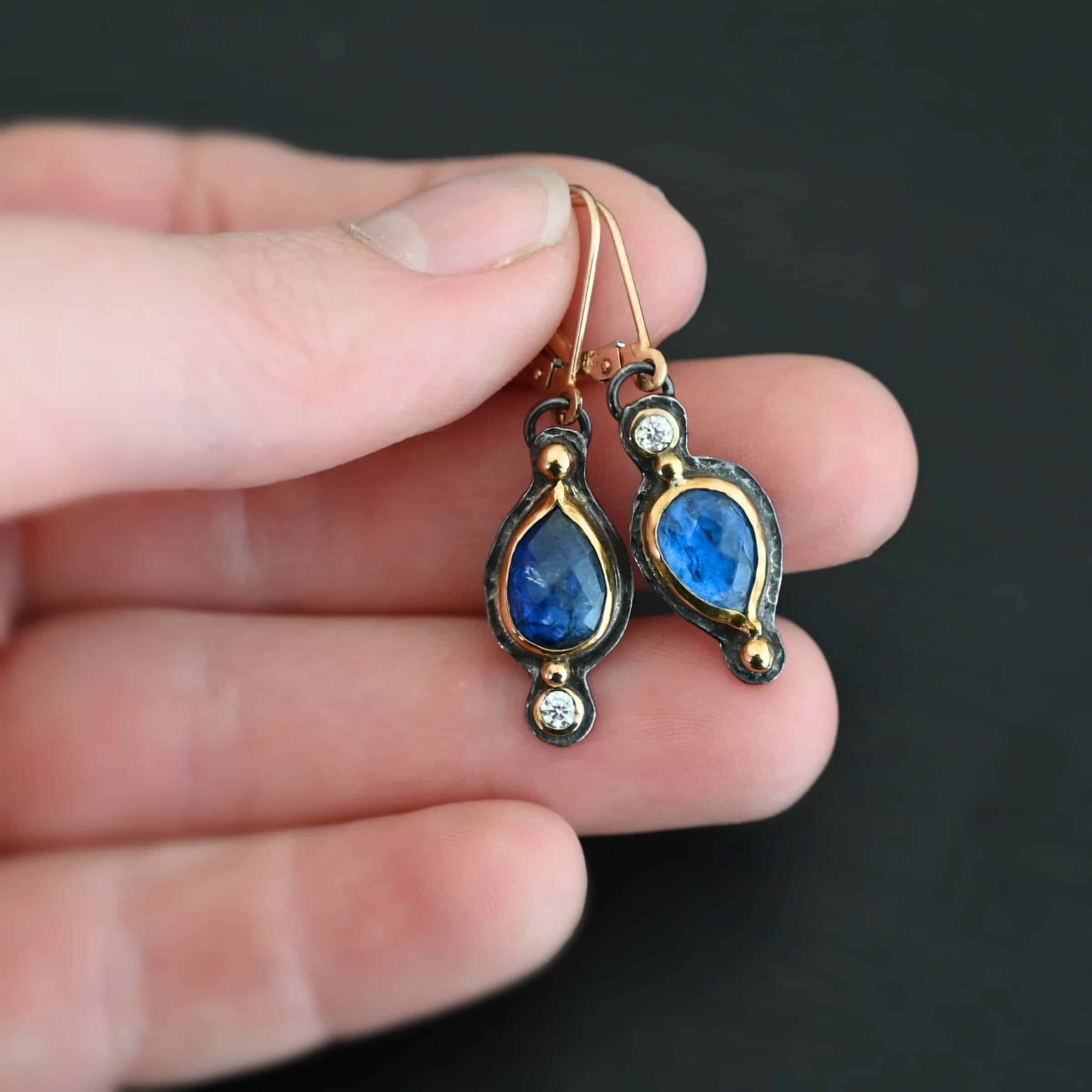 Beyond The Ocean Earrings New Close up of our "Beyond The Ocean" Earrings. Made out of sterling silver and 18k gold, kyanite and moissanites. There are two kyanite stones and two moissanites. Both kyanite stones are teardrops. One is facing with the point upwards. The other has the point facing downwards. Both kyanites are set in gold. The earring with the point facing upwards has a moissanite on the bottom of it. The right earring with the point facing downwards has the moissanite on the top of the earring. Both earrings has two small gold dots on them, one on the top of the earring and one on the bottom. Earrings are on gold leverback ear wires. Earrings are held by a model up close.