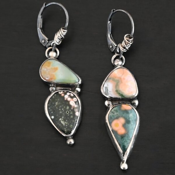 Close up of the "In This Universe" Earrings. Handmade out of sterling silver and ocean jasper. Two stones per each earring. Each earring has four silver dots. One on the top, one on the left side and right side of the earring, and one on the bottom of the earring. Stones vary in shape and size. The left earring has a top stone that is triangle shape and mostly light orange. The bottom stone is mostly green with dark orange dots scattered. The right earring has a top stone that is teardrop shaped, but is placed sideways. It is green with dark brown covering the round end of the teardrop stone. The bottom stone is also teardrop but with the point facing downwards. It is mostly black with pink dots going around the top and right side edge. Earrings are on leverback ear wires with the bottoms of the wires wrapped in sterling silver. Earrings are set against a dark background.
