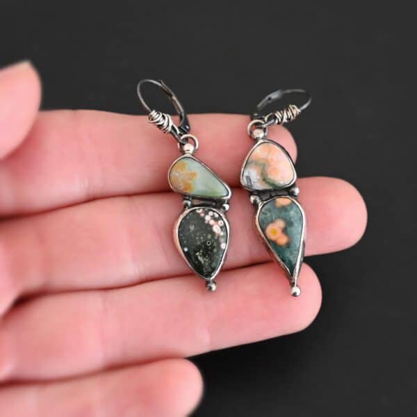 Close up of the "In This Universe" Earrings. Handmade out of sterling silver and ocean jasper. Two stones per each earring. Each earring has four silver dots. One on the top, one on the left side and right side of the earring, and one on the bottom of the earring. Stones vary in shape and size. The left earring has a top stone that is triangle shape and mostly light orange. The bottom stone is mostly green with dark orange dots scattered. The right earring has a top stone that is teardrop shaped, but is placed sideways. It is green with dark brown covering the round end of the teardrop stone. The bottom stone is also teardrop but with the point facing downwards. It is mostly black with pink dots going around the top and right side edge. Earrings are on leverback ear wires with the bottoms of the wires wrapped in sterling silver. Earrings are set against the fingertips of a hand.