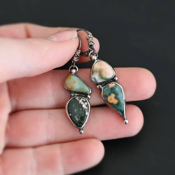 Close up of the "In This Universe" Earrings. Handmade out of sterling silver and ocean jasper. Two stones per each earring. Each earring has four silver dots. One on the top, one on the left side and right side of the earring, and one on the bottom of the earring. Stones vary in shape and size. The left earring has a top stone that is triangle shape and mostly light orange. The bottom stone is mostly green with dark orange dots scattered. The right earring has a top stone that is teardrop shaped, but is placed sideways. It is green with dark brown covering the round end of the teardrop stone. The bottom stone is also teardrop but with the point facing downwards. It is mostly black with pink dots going around the top and right side edge. Earrings are on leverback ear wires with the bottoms of the wires wrapped in sterling silver. Earrings are set against the fingertips of a hand.