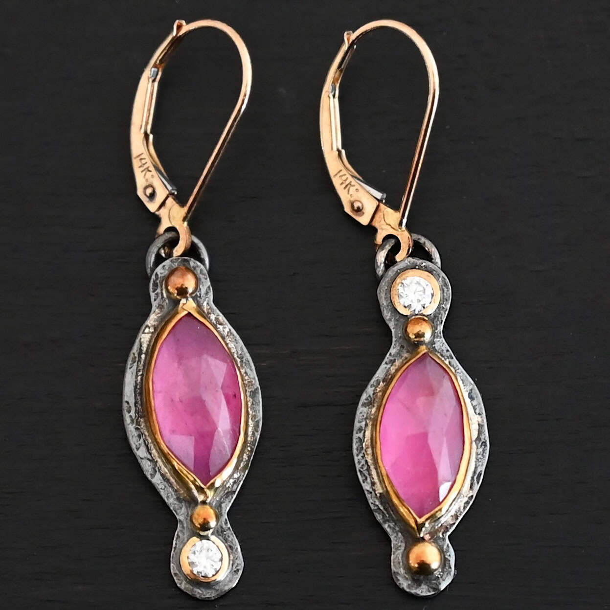 Close up of our "Pink Skies" Earrings. Made out of sterling silver and 18k gold, rubies and moissanites. There are two ruby stones and two moissanites. Both ruby stones are oval shaped with points on each end. Both rubies are set in gold. The left earring has a moissanite set in gold on the bottom of it. The right earring has the moissanite on the top of the earring. Both earrings have two small gold dots on them, one on the top of the earring and one on the bottom. Earrings are on gold leverback ear wires.