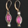 Close up of our "Pink Skies" Earrings. Made out of sterling silver and 18k gold, rubies and moissanites. There are two ruby stones and two moissanites. Both ruby stones are oval shaped with points on each end. Both rubies are set in gold. The left earring has a moissanite set in gold on the bottom of it. The right earring has the moissanite on the top of the earring. Both earrings have two small gold dots on them, one on the top of the earring and one on the bottom. Earrings are on gold leverback ear wires.