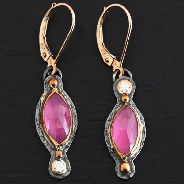 Close up of our "Pink Skies" Earrings. Made out of sterling silver and 18k gold, rubies and moissanites. There are two ruby stones and two moissanites. Both ruby stones are oval shaped with points on each end. Both rubies are set in gold. The left earring has a moissanite set in gold on the bottom of it. The right earring has the moissanite on the top of the earring. Both earrings have two small gold dots on them, one on the top of the earring and one on the bottom. Earrings are on gold leverback ear wires.