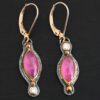 Close up of our "Pink Skies" Earrings. Made out of sterling silver and 18k gold, rubies and moissanites. There are two ruby stones and two moissanites. Both ruby stones are oval shaped with points on each end. Both rubies are set in gold. The left earring has a moissanite set in gold on the bottom of it. The right earring has the moissanite on the top of the earring. Both earrings have two small gold dots on them, one on the top of the earring and one on the bottom. Earrings are on gold leverback ear wires.