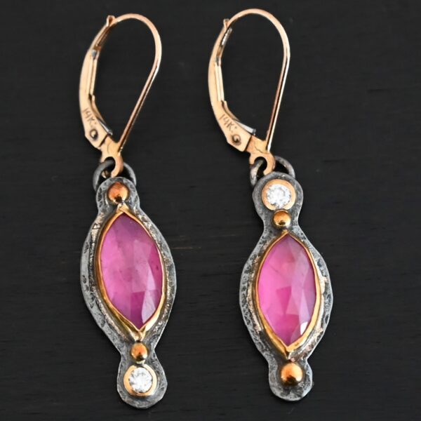 Close up of our "Pink Skies" Earrings. Made out of sterling silver and 18k gold, rubies and moissanites. There are two ruby stones and two moissanites. Both ruby stones are oval shaped with points on each end. Both rubies are set in gold. The left earring has a moissanite set in gold on the bottom of it. The right earring has the moissanite on the top of the earring. Both earrings have two small gold dots on them, one on the top of the earring and one on the bottom. Earrings are on gold leverback ear wires.