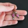 Close up of our "Pink Skies" Earrings. Made out of sterling silver and 18k gold, rubies and moissanites. There are two ruby stones and two moissanites. Both ruby stones are oval shaped with points on each end. Both rubies are set in gold. The left earring has a moissanite set in gold on the bottom of it. The right earring has the moissanite on the top of the earring. Both earrings have two small gold dots on them, one on the top of the earring and one on the bottom. Earrings are on gold leverback ear wires. Earrings are held by a model up close.