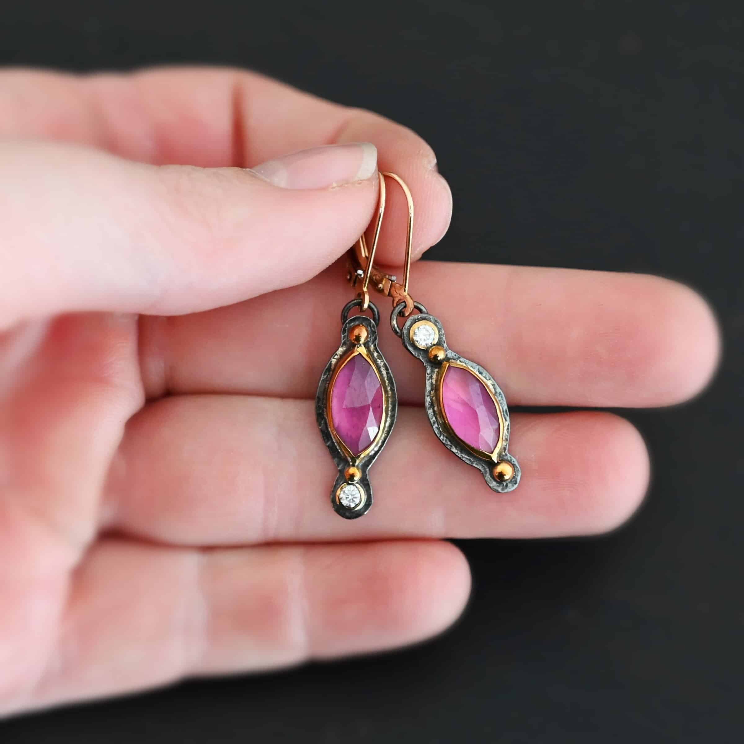 Close up of our "Pink Skies" Earrings. Made out of sterling silver and 18k gold, rubies and moissanites. There are two ruby stones and two moissanites. Both ruby stones are oval shaped with points on each end. Both rubies are set in gold. The left earring has a moissanite set in gold on the bottom of it. The right earring has the moissanite on the top of the earring. Both earrings have two small gold dots on them, one on the top of the earring and one on the bottom. Earrings are on gold leverback ear wires. Earrings are held by a model up close.