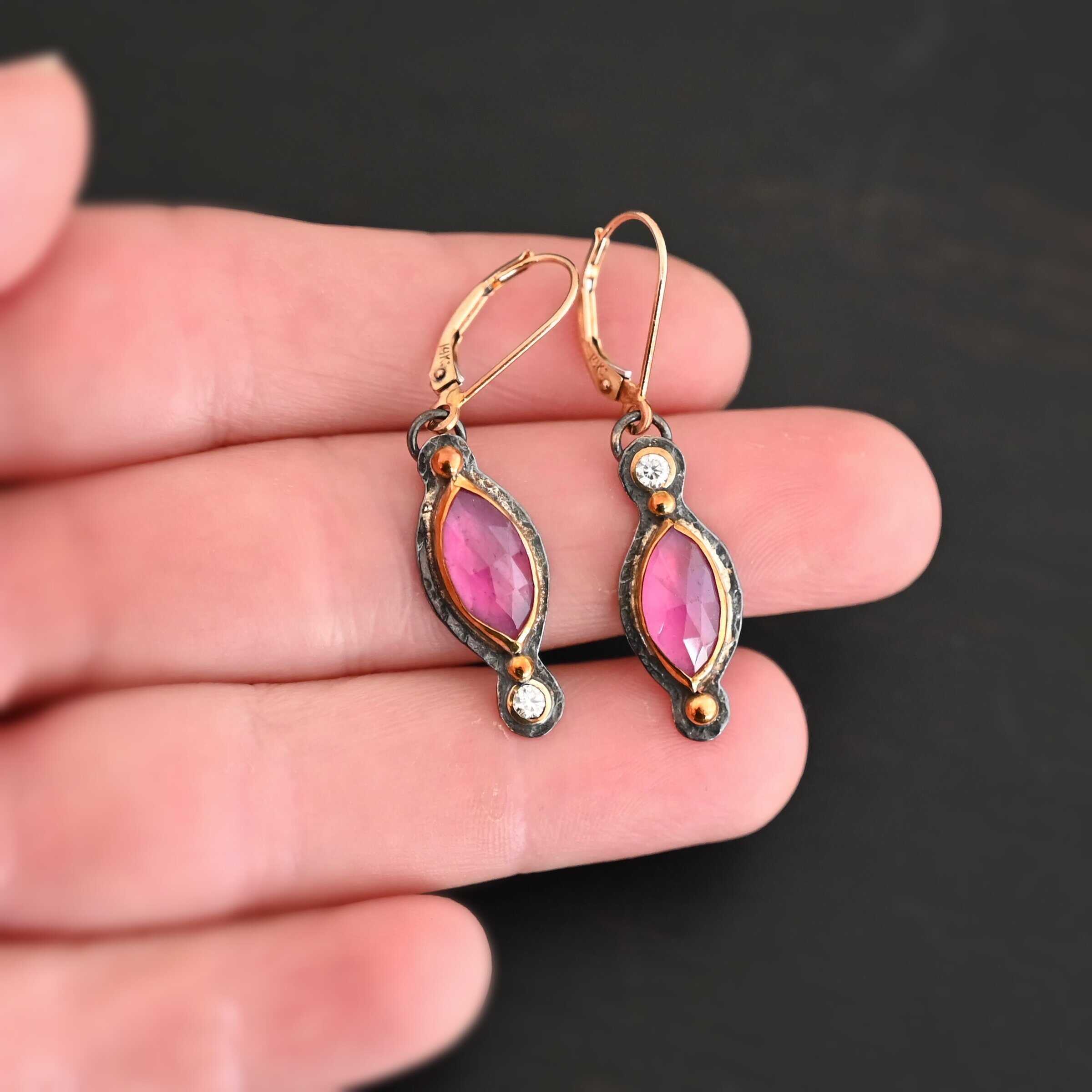 Close up of our "Pink Skies" Earrings. Made out of sterling silver and 18k gold, rubies and moissanites. There are two ruby stones and two moissanites. Both ruby stones are oval shaped with points on each end. Both rubies are set in gold. The left earring has a moissanite set in gold on the bottom of it. The right earring has the moissanite on the top of the earring. Both earrings have two small gold dots on them, one on the top of the earring and one on the bottom. Earrings are on gold leverback ear wires. Earrings are held by a model up close.