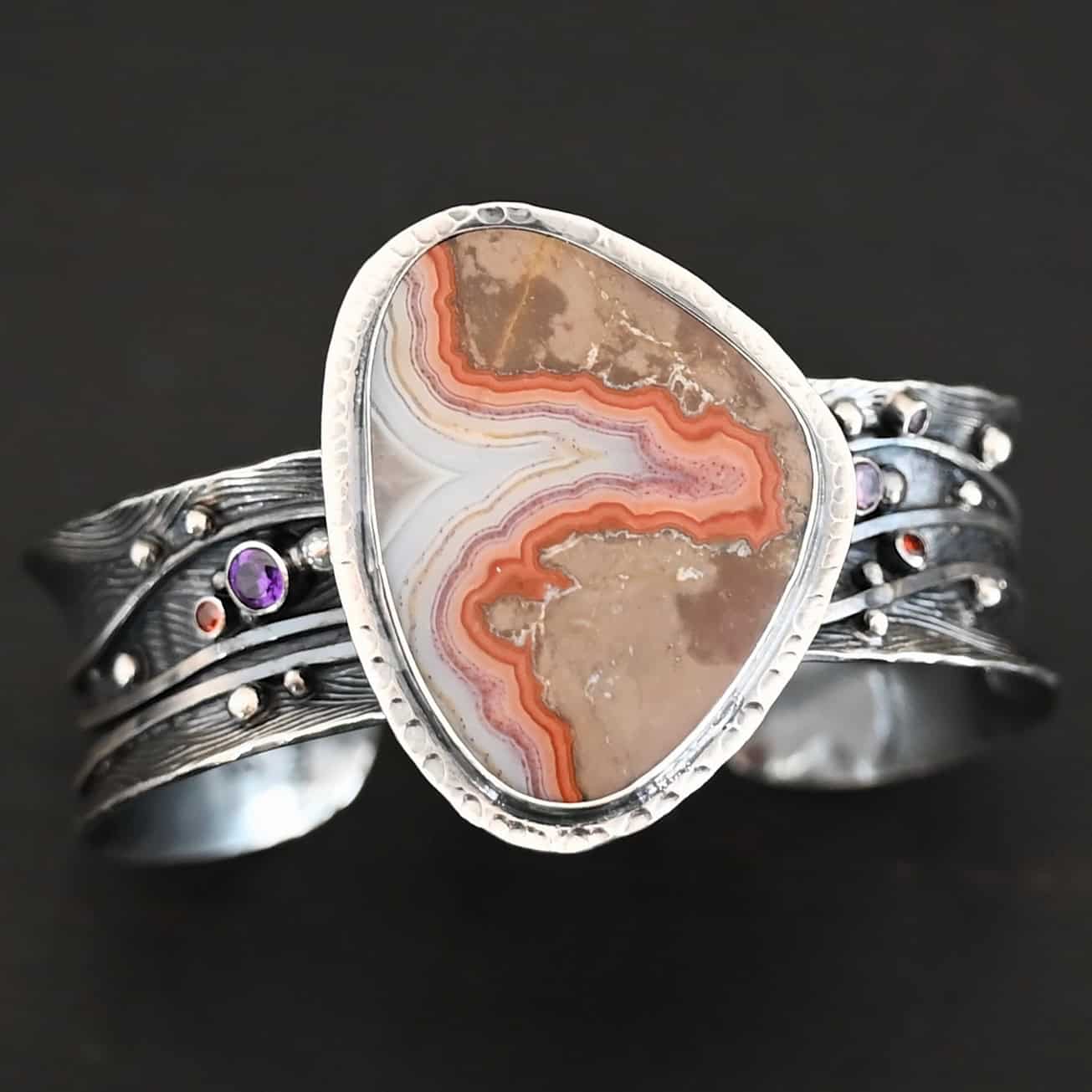 Zion Canyon Cuff "Zion Canyon" Cuff. There is a large, wide, oval, vertical ocean jasper in the center of a very wide, thick cuff. There are wavy lines of silver going across it and multiple gemstones scattered across it, along with silver ball detail. There is hammered silver around the stone.