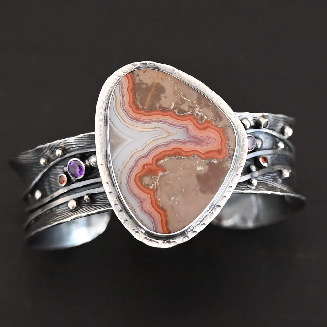 Zion Canyon Cuff "Zion Canyon" Cuff. There is a large, wide, oval, vertical ocean jasper in the center of a very wide, thick cuff. There are wavy lines of silver going across it and multiple gemstones scattered across it, along with silver ball detail. There is hammered silver around the stone.