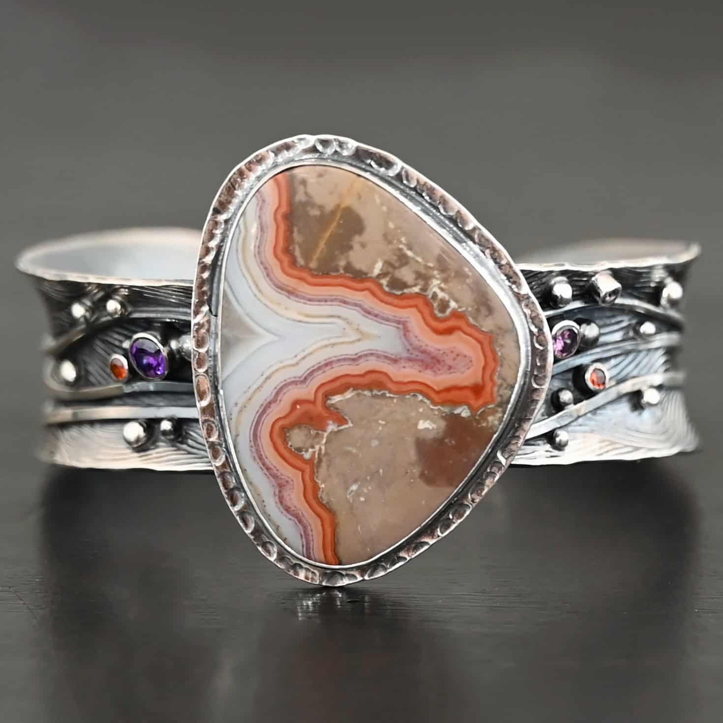 Zion Canyon Cuff "Zion Canyon" Cuff. There is a large, wide, oval, vertical ocean jasper in the center of a very wide, thick cuff. There are wavy lines of silver going across it and multiple gemstones scattered across it, along with silver ball detail. There is hammered silver around the stone.