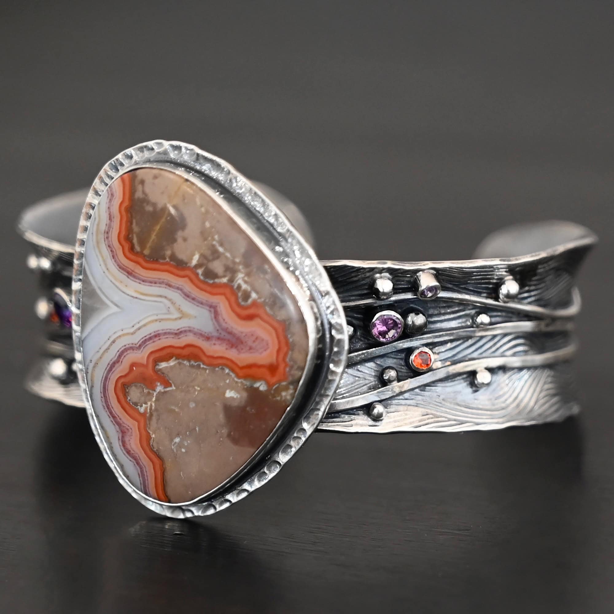 Zion Canyon Cuff "Zion Canyon" Cuff. There is a large, wide, oval, vertical ocean jasper in the center of a very wide, thick cuff. There are wavy lines of silver going across it and multiple gemstones scattered across it, along with silver ball detail. There is hammered silver around the stone.